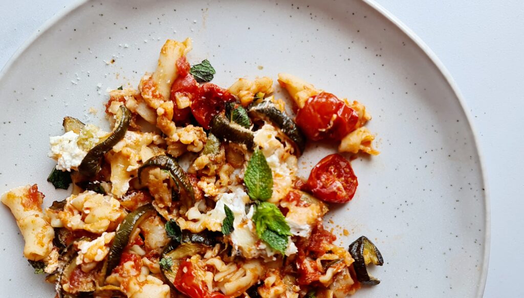 A close-up of a plate of tomato-based zucchini pasta bake, garnished with cherry tomatoes and crumbled cheese. Fresh mint leaves are scattered on top, adding a pop of green color. The dish is presented on a light-colored plate.