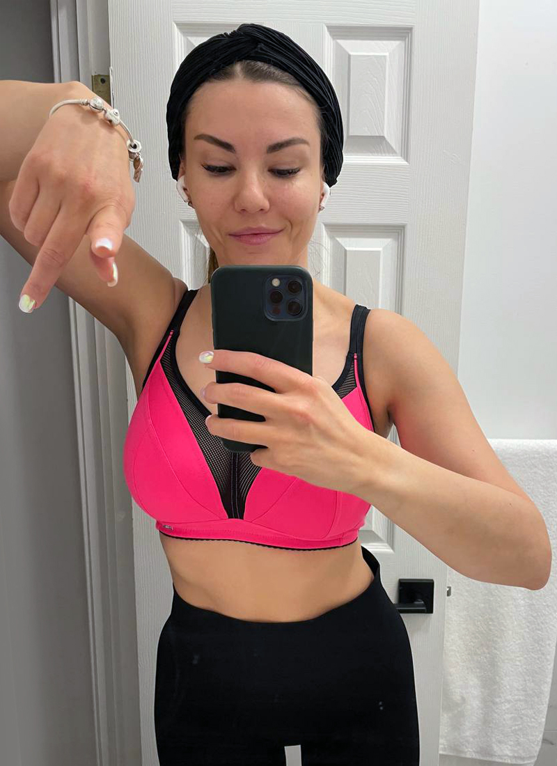 A person wearing a black headband, pink sports bra, and black leggings takes a selfie in front of a bathroom mirror. Pointing downwards with their left hand and holding the phone with their right, they showcase their dedication to fitness goals. The bathroom features a white towel hanging on a rack.