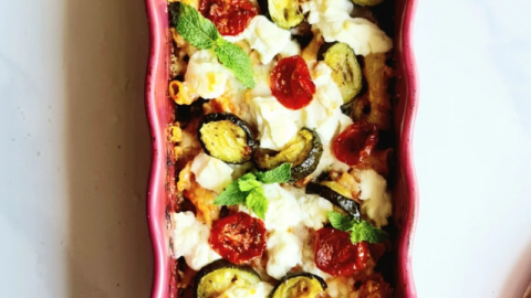 A rectangular red baking dish filled with a colorful zucchini tomato pasta bake. The dish contains rigatoni pasta, zucchini slices, cherry tomatoes, ricotta cheese, and is garnished with fresh mint leaves. The background shows a white surface with a small wooden board and a glass container.