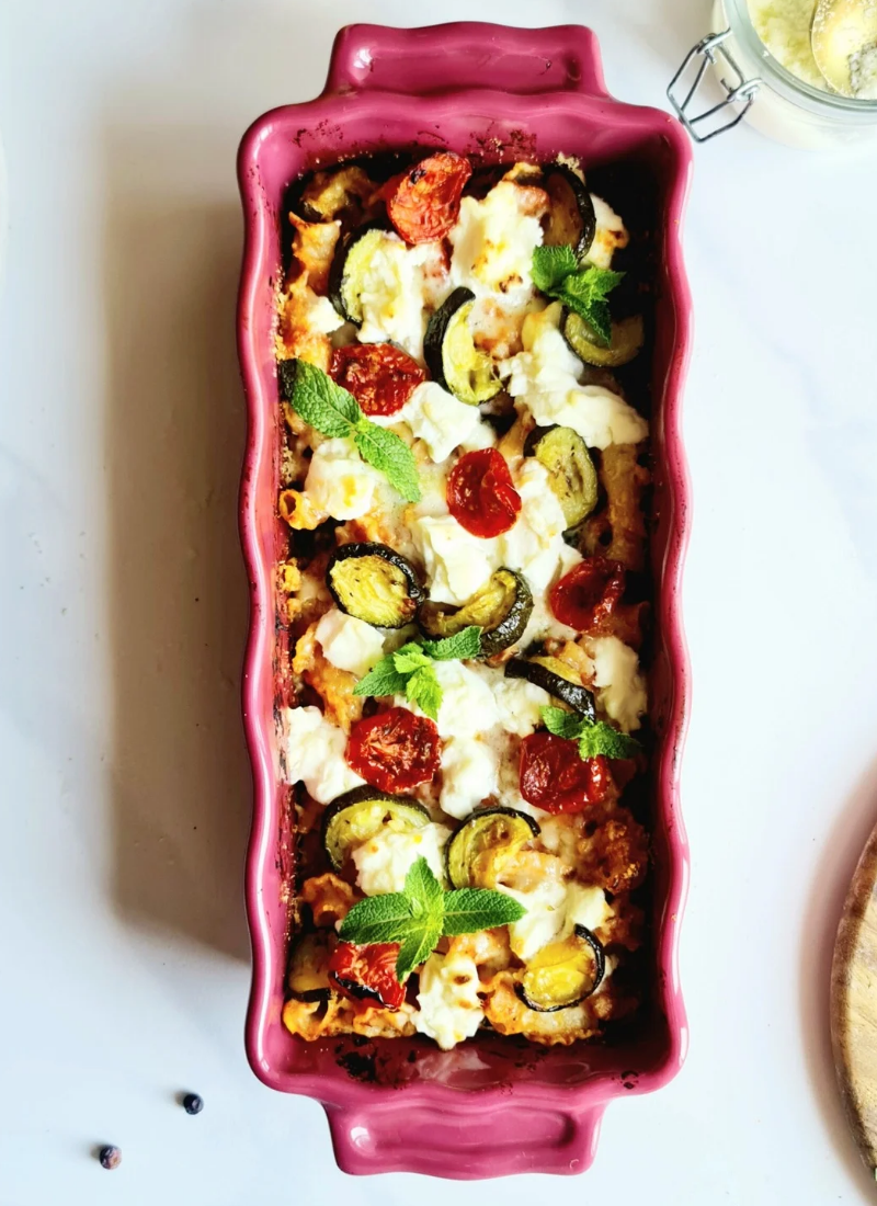 A rectangular red baking dish filled with a colorful zucchini tomato pasta bake. The dish contains rigatoni pasta, zucchini slices, cherry tomatoes, ricotta cheese, and is garnished with fresh mint leaves. The background shows a white surface with a small wooden board and a glass container.