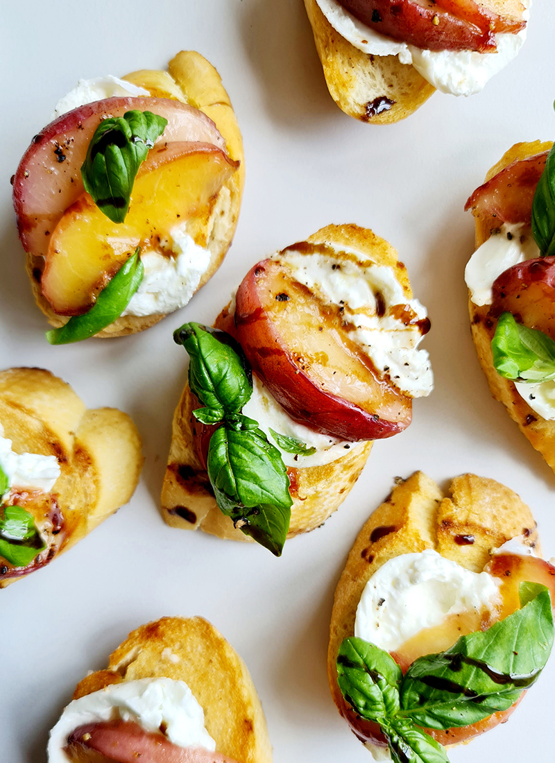 Close-up of peach bruschetta crostini topped with whipped burrata, thin slices of peaches, cracked black pepper, and a fresh basil leaf. The background is a plain white surface, giving focus to the colorful and appetizing toppings.