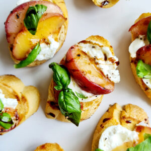 Close-up ofpeach bruschetta crostini topped with whipped burrata, thin slices of peaches, cracked black pepper, and a fresh basil leaf. The background is a plain white surface, giving focus to the colorful and appetizing toppings.