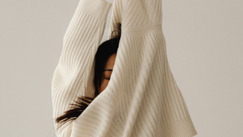 Person in a beige ribbed sweater with long sleeves, stretching their arms upwards to relieve stress naturally, so their face is partially obscured by the garment. The background is a plain, neutral tone.