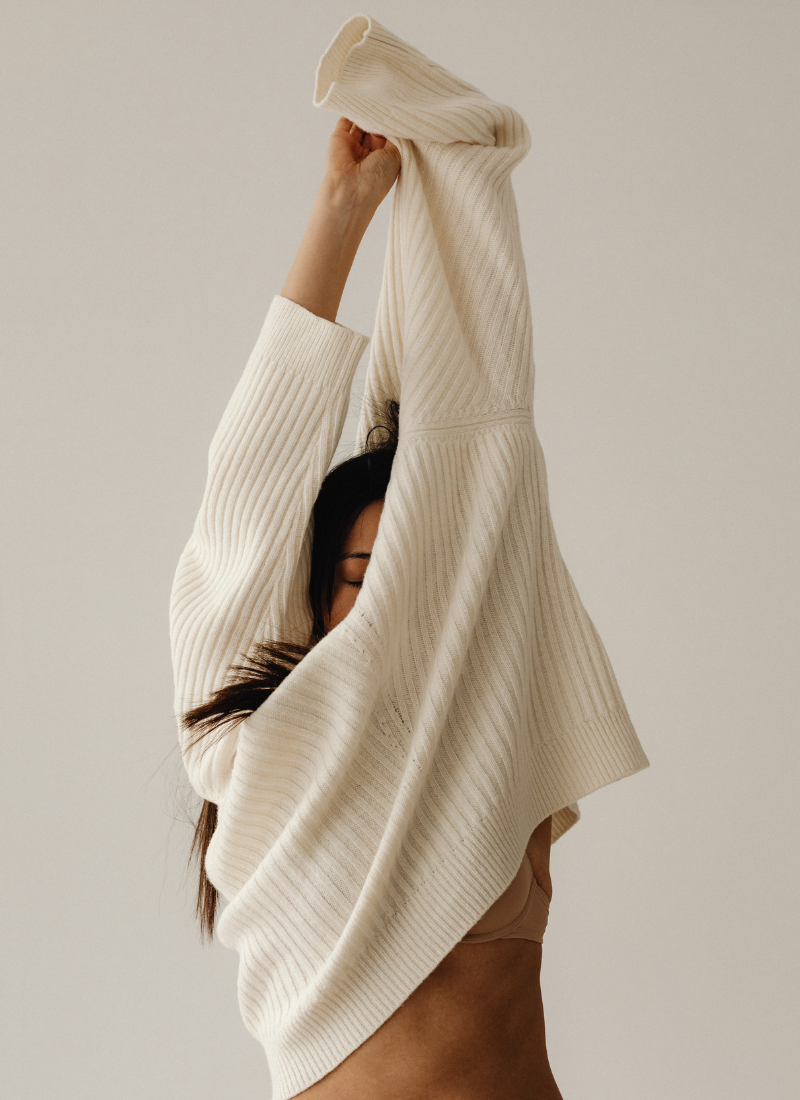 Person in a beige ribbed sweater with long sleeves, stretching their arms upwards to relieve stress naturally, so their face is partially obscured by the garment. The background is a plain, neutral tone.