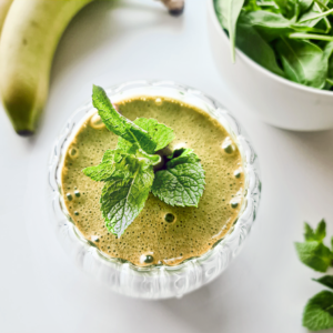 A glass of Cindy Crawford’s green smoothie garnished with a sprig of fresh mint. A banana and a bowl of spinach leaves are in the background, all placed on a white surface.