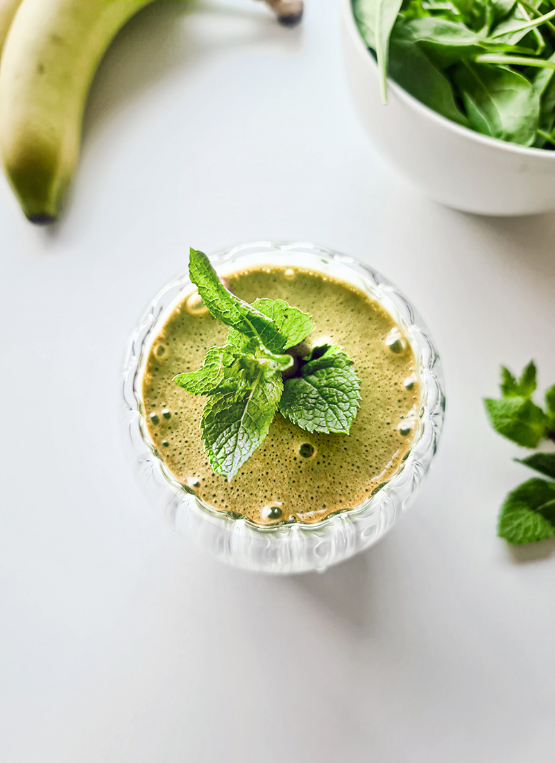 A green smoothie garnished with fresh mint leaves sits in a glass on a white surface. A banana and a bowl of spinach leaves are visible in the background. This model smoothie, reminiscent of Cindy Crawford’s green smoothie, boasts a bright and fresh presentation with healthy ingredients.