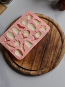 A pink silicone Halloween mold filled with baked mozzarella sits on a round wooden cutting board. The mold features various shapes. This quirky scene on a light-colored countertop is perfect for crafting spooky Halloween appetizers.