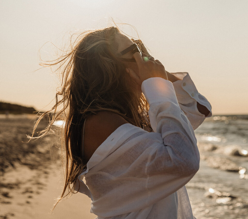 A person wears a white shirt off their shoulders, and the wind blows through their hair. The ocean waves and distant shoreline are visible under the warm, golden light, reminding her how to develop a positive mindset amidst negative self-talk.