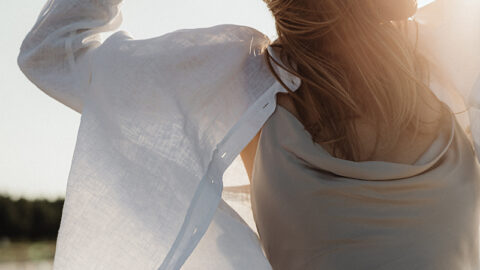 A woman stands on a beach, her arms raised and wearing sunglasses. Dressed in a light dress and a white button-down shirt, she embodies the essence of how to develop a positive mindset. Her hair is blowing in the wind and the sunlight creates a warm glow around her, with a scenic view of sand and trees in the background.