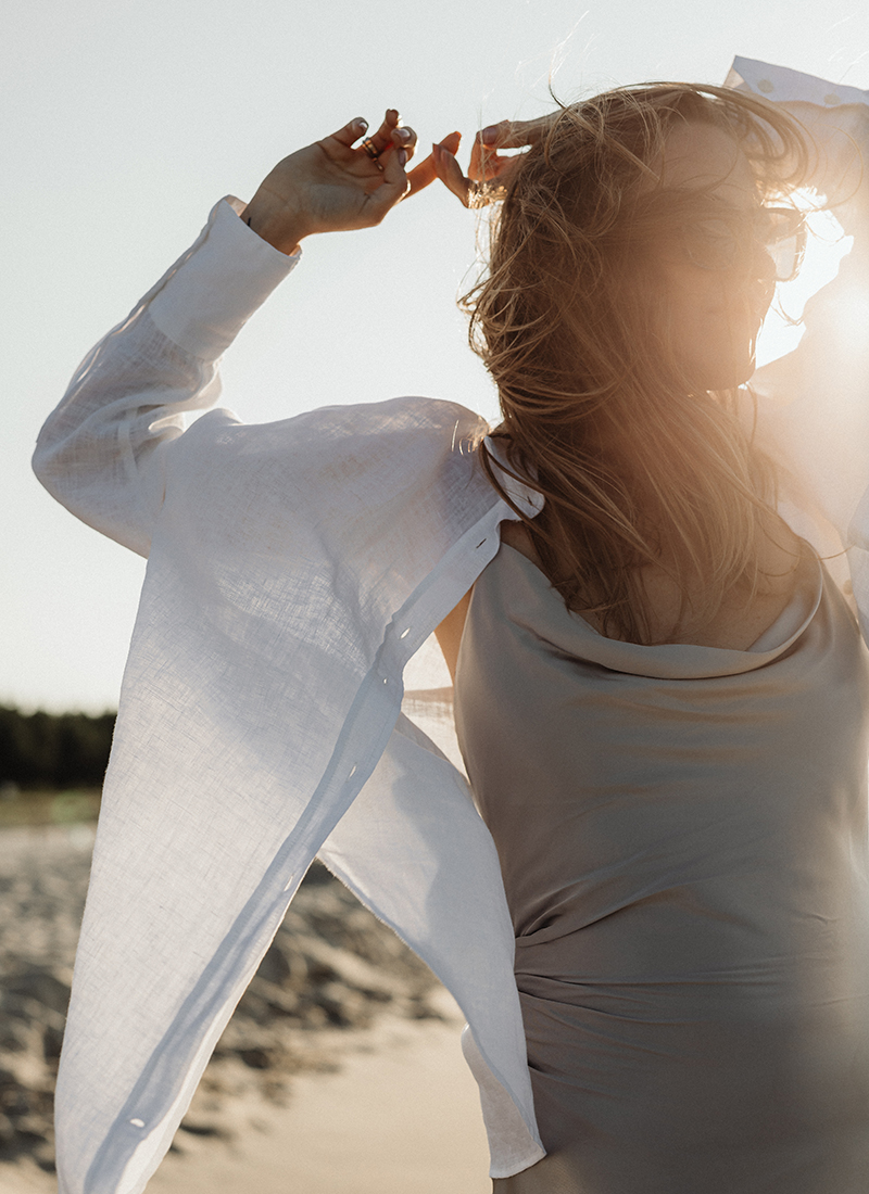 A woman stands on a beach, her arms raised and wearing sunglasses. Dressed in a light dress and a white button-down shirt, she embodies the essence of how to develop a positive mindset. Her hair is blowing in the wind and the sunlight creates a warm glow around her, with a scenic view of sand and trees in the background.