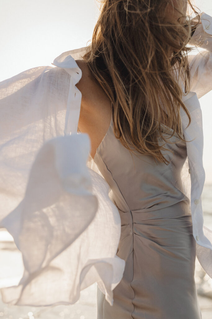 A woman stands on a beach, with her back toward the camera. She wears a silky dress and a flowing, lightweight white shirt that catches the breeze. Her long hair is tousled by the wind, embodying the freedom one can feel when learning how to develop a positive mindset.