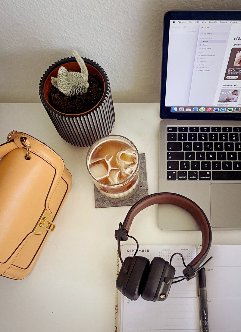 A workspace featuring a laptop with a document open, a planner showing the month of September, a black pair of headphones for streaming motivational podcasts, a beige handbag, a potted cactus, and a glass of iced coffee on a coaster.