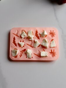 A pink silicone mold designed with various Halloween-themed shapes. Some white, mozzarella pieces are partially filling several of the molds. The mold is placed on a light-colored surface, perfect for creating treats or even spooky Halloween appetizers.