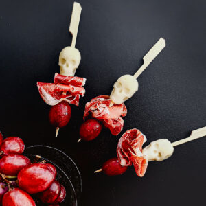 Three wooden Halloween mozzarella skewers, each adorned with a small white skull decoration, slices of cured meat, and two red grapes, are arranged on a dark surface. These spooky Halloween appetizers make for an eerie yet delicious treat. In the bottom left corner, more red grapes are visible in a clear bowl.