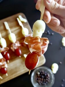 A hand holds a skewer with a grape, a piece of prosciutto, and a cheese skull - a spooky Halloween appetizer idea. In the background, a wooden board displays several similar mozzarella skewers, amidst scattered grapes and peppercorns on a black surface. A bowl with more peppercorns is also visible.