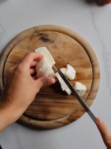 A hand holding and cutting a block of white cheese on a round wooden cutting board with a knife. The background is a light grey countertop, perfect for creating spooky Halloween appetizers with mozzarella