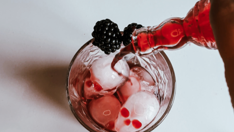 A hand is pouring a red liquid from a glass bottle into a glass filled with ice cubes, creating a festive Blackberry Aperol Spritz. Garnished with a blackberry, the scene is set with other blackberries and a lit candle in the background on the white surface—perfect for a Halloween cocktail delight.