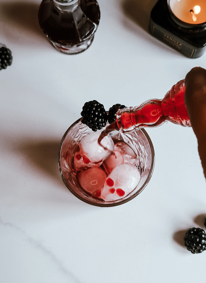 A hand is pouring a red liquid from a glass bottle into a glass filled with ice cubes, creating a festive Blackberry Aperol Spritz. Garnished with a blackberry, the scene is set with other blackberries and a lit candle in the background on the white surface—perfect for a Halloween cocktail delight.