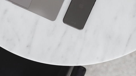 A minimalist workspace balancing work and life features a silver laptop and a dark smartphone on a round white marble table. A brown coat is draped over a black chair beside the table, creating a clean and stylish setting that promotes mental well-being.