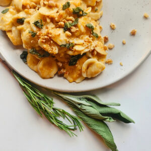 A plate of pumpkin pasta in an orange sauce is topped with chopped herbs and crumbled walnuts. Fresh rosemary and sage sprigs lie beside the plate on a pristine white surface, adding a touch of elegance to the dish.