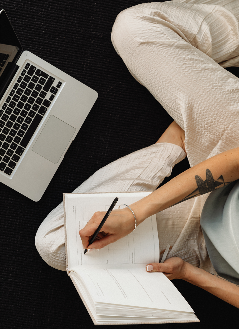 A person sits cross-legged on a dark carpet, writing in an open notebook with a pen—a mindful practice to enhance focus. A laptop rests nearby to their left. They wear light-colored pants, and a bracelet, with a tattoo visible on their right arm, embodying mindfulness for women working from home.