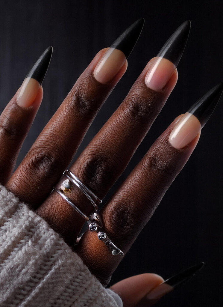 A hand with long, black-tipped nails, featuring an intricate Halloween nail design, is adorned with silver rings. The person is wearing a white knitted sweater. A dark background enhances the focus on the hand and eerie Halloween manicure.