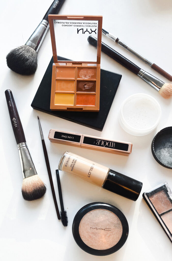 A flat lay of various makeup products and brushes on a white background, perfect for women working from home aiming to look good on video. Items include foundation, powder, eyeshadow, eyeliner, and a mascara wand alongside several brushes, all arranged in a scattered manner.