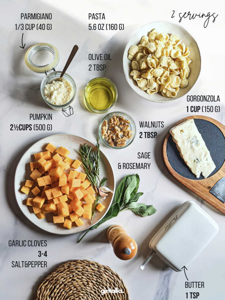 Displayed elegantly are the ingredients for a risotto-style pumpkin pasta dish: grated Parmigiano, orecchiette, olive oil, diced pumpkin, gorgonzola cheese, walnuts, sage and rosemary sprigs, garlic cloves, and butter with salt and pepper. Each ingredient specifies its quantity.