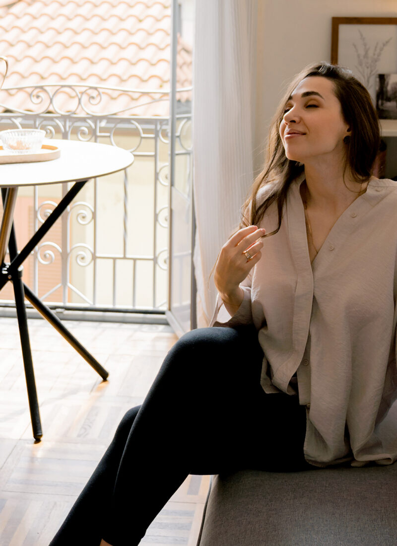 A woman sits on a gray couch in a bright room with a round table and an open balcony door, practicing gratitude. She wears a beige shirt and black pants, appearing relaxed with her eyes closed and a hand near her hair. A framed picture and lamp adorn the shelf behind her.